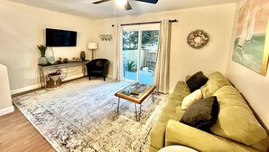 Ein Wohnzimmer mit Deckenventilator, Fernseher, einer Couch, einem Couchtisch und einem großen Fenster mit Vorhängen.