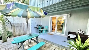 Eine Holzterrasse mit Picknicktischen und Grill, ein überdachter Patio-Bereich und eine Glastür, die zu einem Innenraum führt.