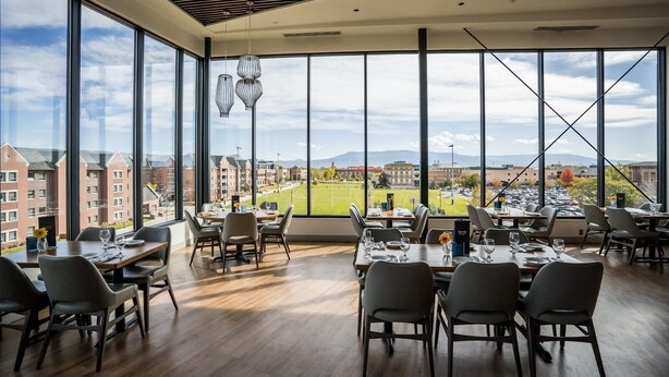 Un espace repas doté de grandes fenêtres donnant sur un terrain de sport et des bâtiments. Des tables sont disposées, accompagnées de chaises et de couverts.