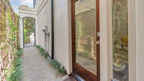 Ein schmaler Weg führt zu einer Tür mit Glasscheibe, begrenzt von einem Holzzaun und einer weißen Mauer.