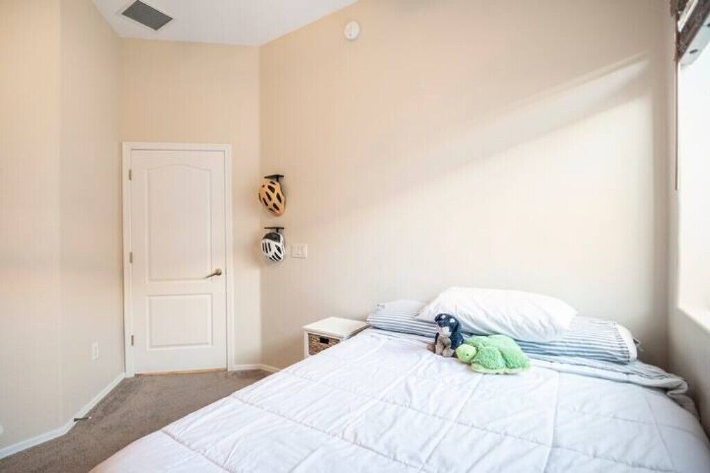 A bedroom with a bed, a door, a small bedside table, and a wall-mounted shelf with a helmet and a small figurine.