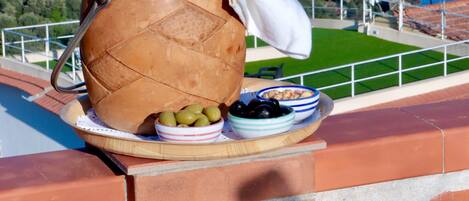 A bottle of wine on a table with olives and a bowl of grapes, overlooking a coastal view with buildings and greenery.