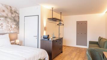 A modern hotel room with a bed, a desk with a lamp, a chair, a sofa, and a wooden floor.