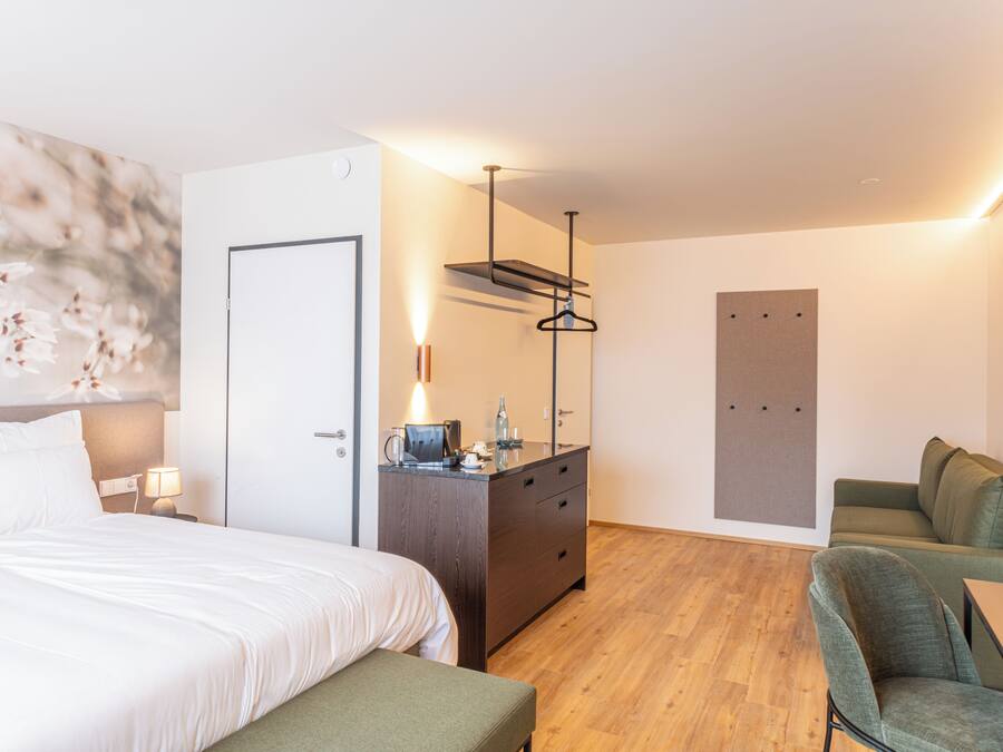 A modern hotel room with a bed, a desk with a lamp, a chair, a sofa, and a wooden floor.