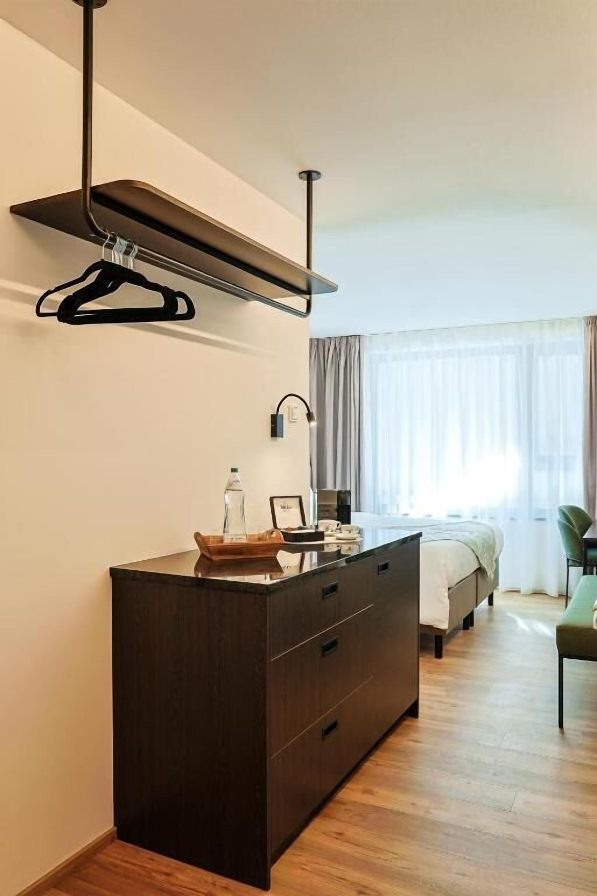 Una habitación de hotel moderna con un aparador de madera oscura, una lámpara colgante, una cama con una silla verde y un ventanal con cortinas.