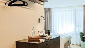 Una habitación de hotel moderna con un aparador de madera oscura, una lámpara colgante, una cama con una silla verde y un ventanal con cortinas.