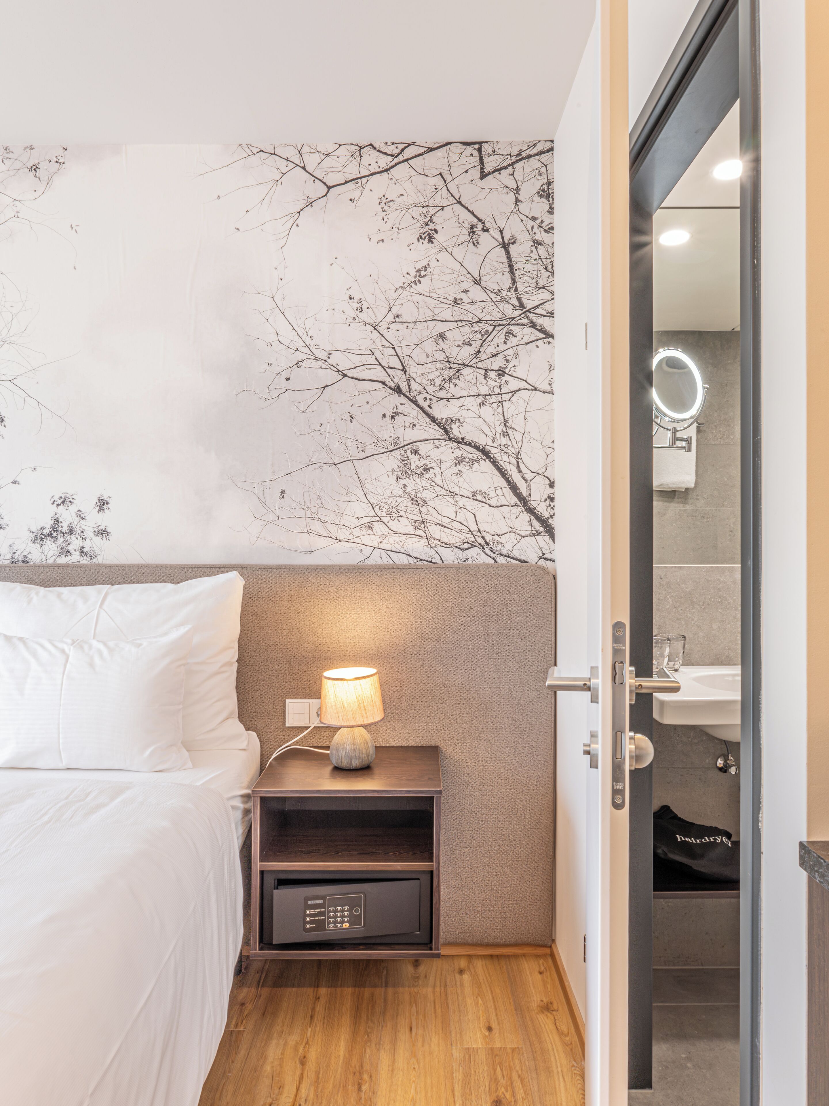 A bedroom with a bed, a nightstand with a lamp, a wall with a tree mural, and an open door to a bathroom.