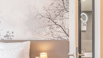 A bedroom with a bed, a nightstand with a lamp, a wall with a tree mural, and an open door to a bathroom.