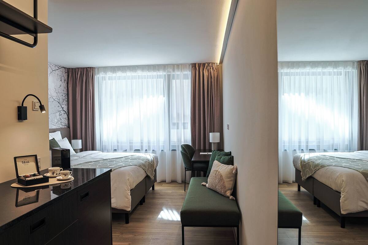 A hotel room with two beds, a desk, a chair, a bench, and a minibar.