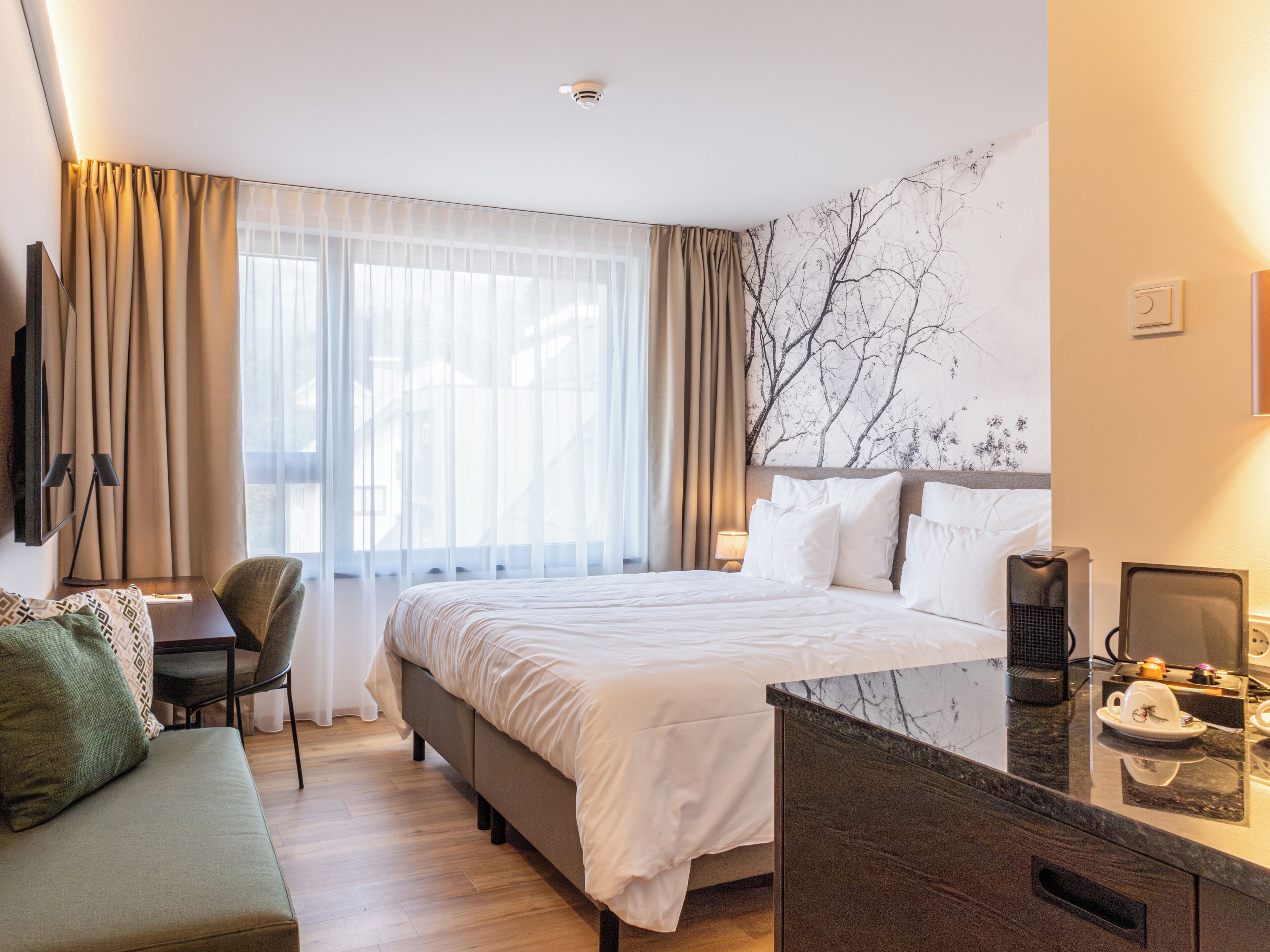 A hotel room with a bed, a desk, a chair, a sofa, and a marble countertop with a coffee maker.