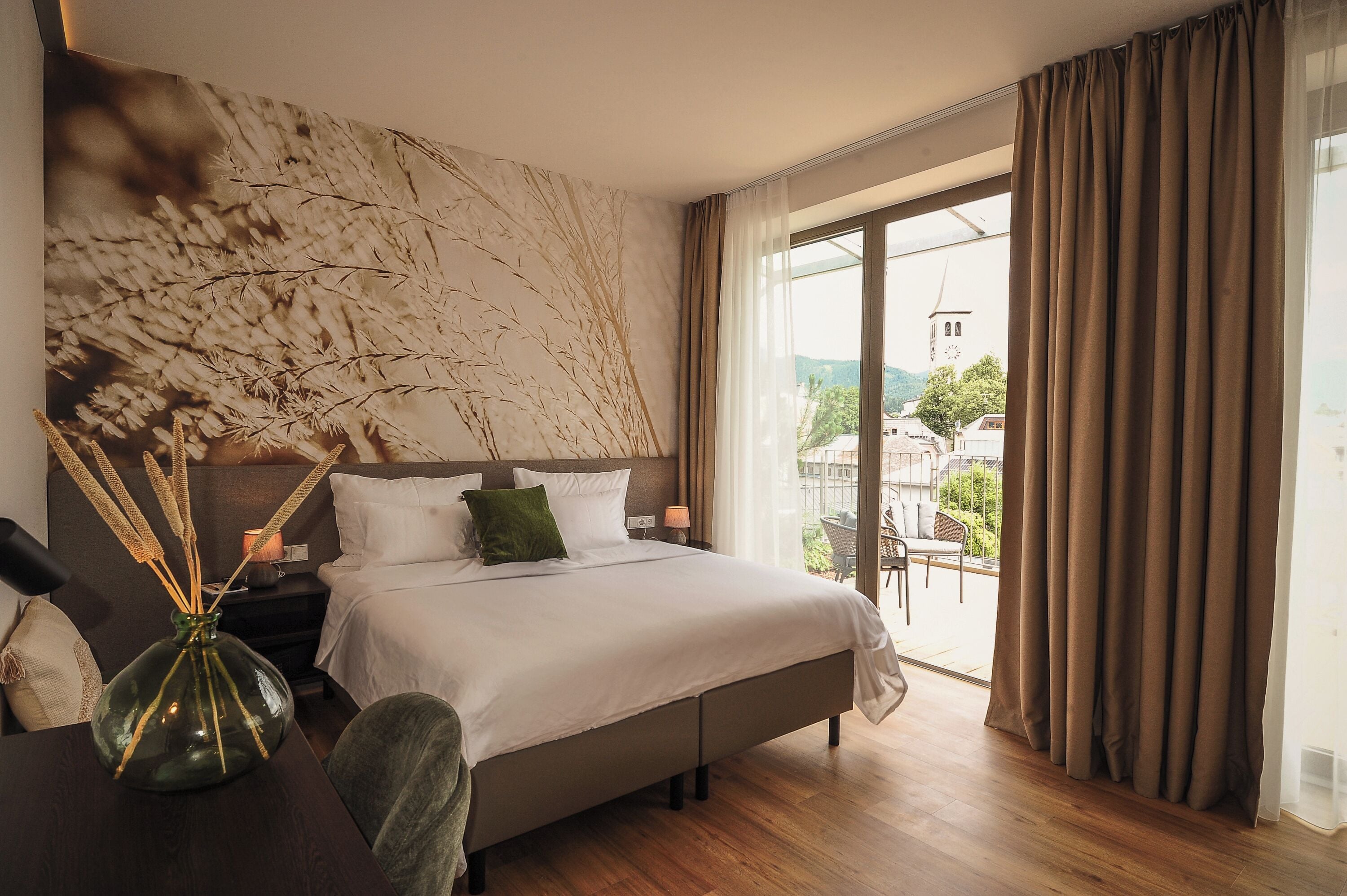 A hotel room with a large bed, a nightstand, a vase with dried plants, and a view of a cityscape through the window.