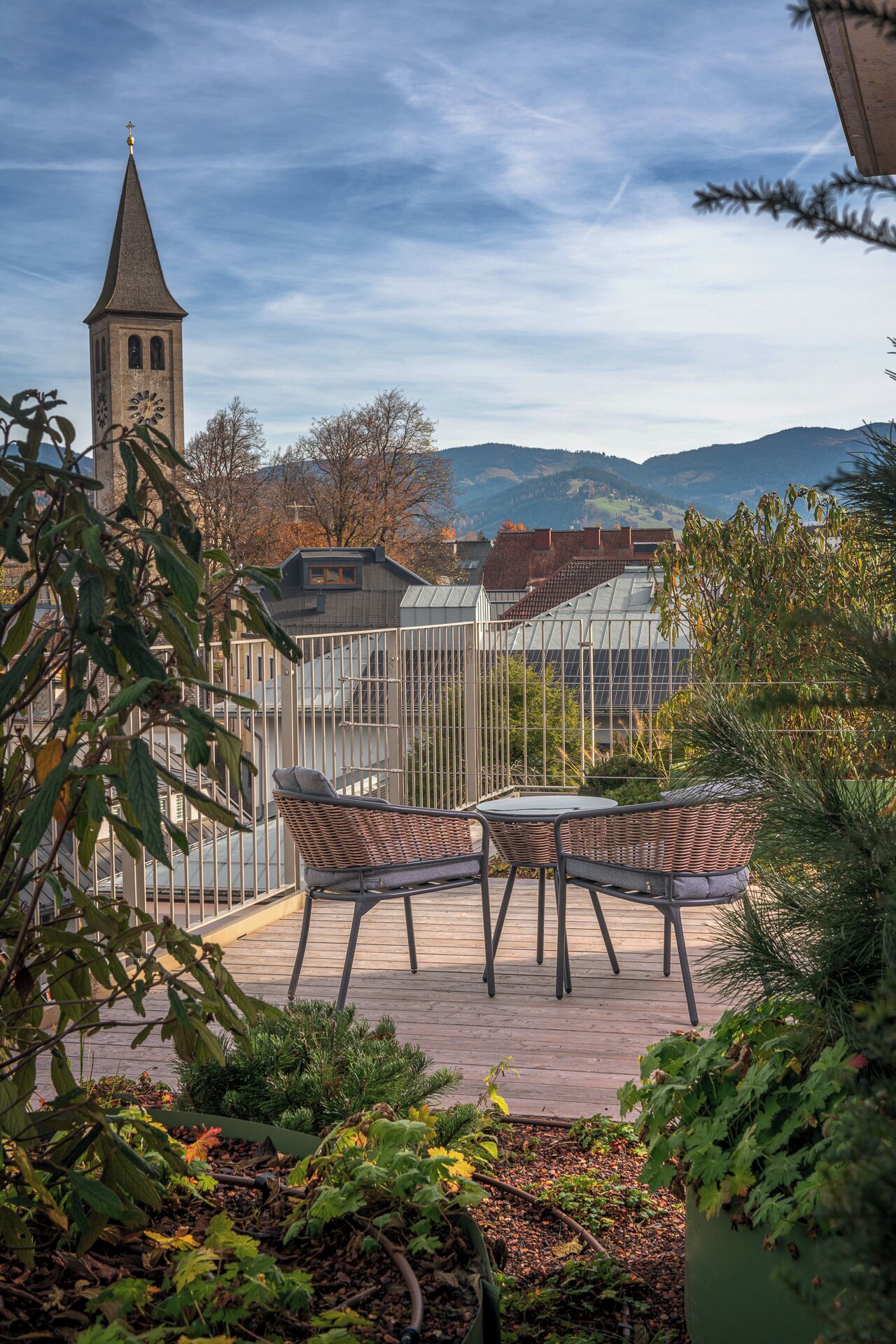 Eine Terrasse mit Korbstühlen und einem Tisch, mit Blick auf einen Kirchturm und eine gebirgige Landschaft.