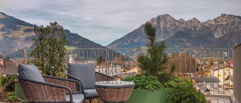 A deck with wicker chairs and a small table, overlooking a mountainous landscape with buildings in the valley.