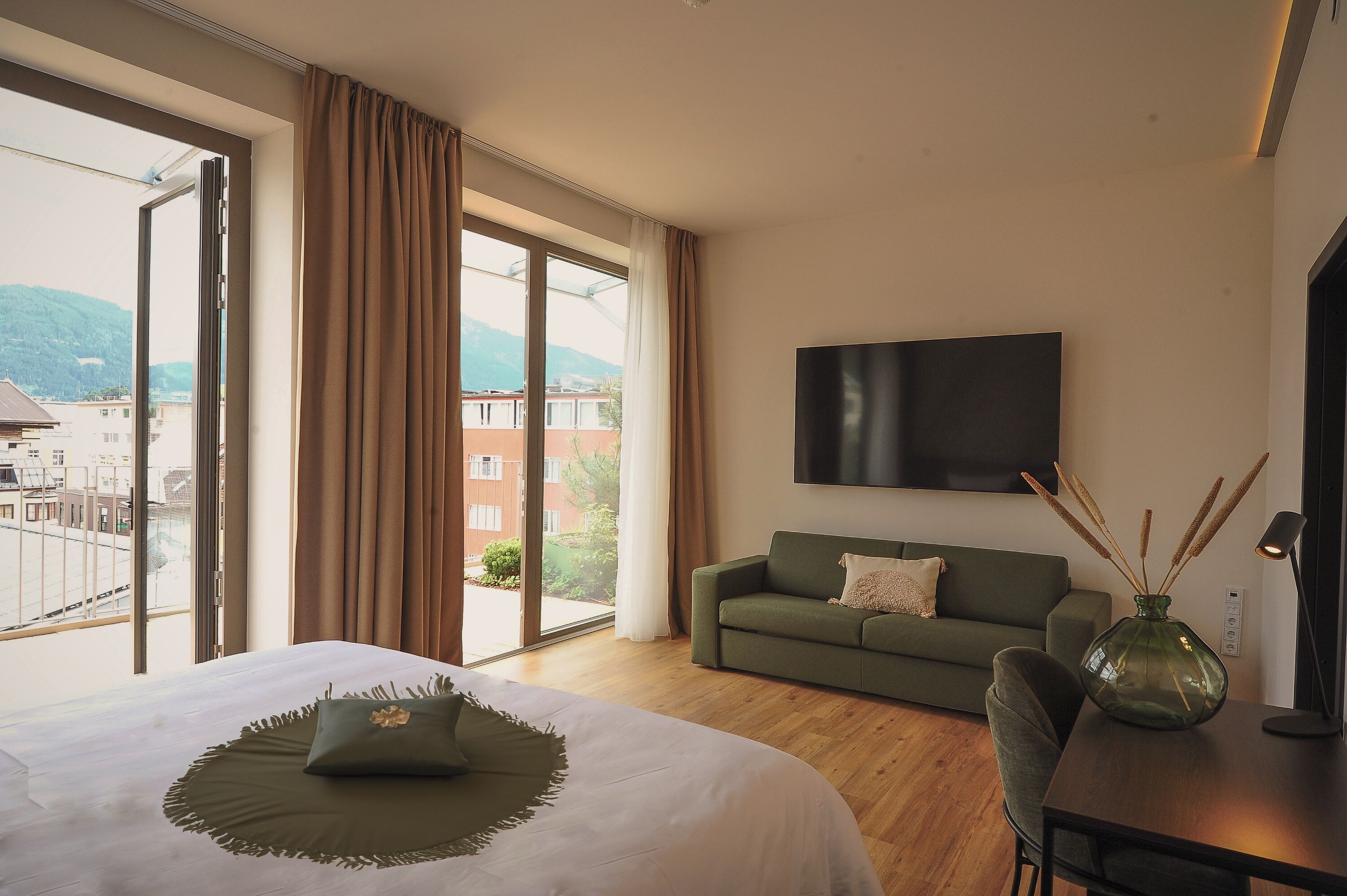 Habitación de hotel con una cama grande, un sofá verde, un televisor de pantalla plana y vistas a edificios y montañas a través de un ventanal amplio.