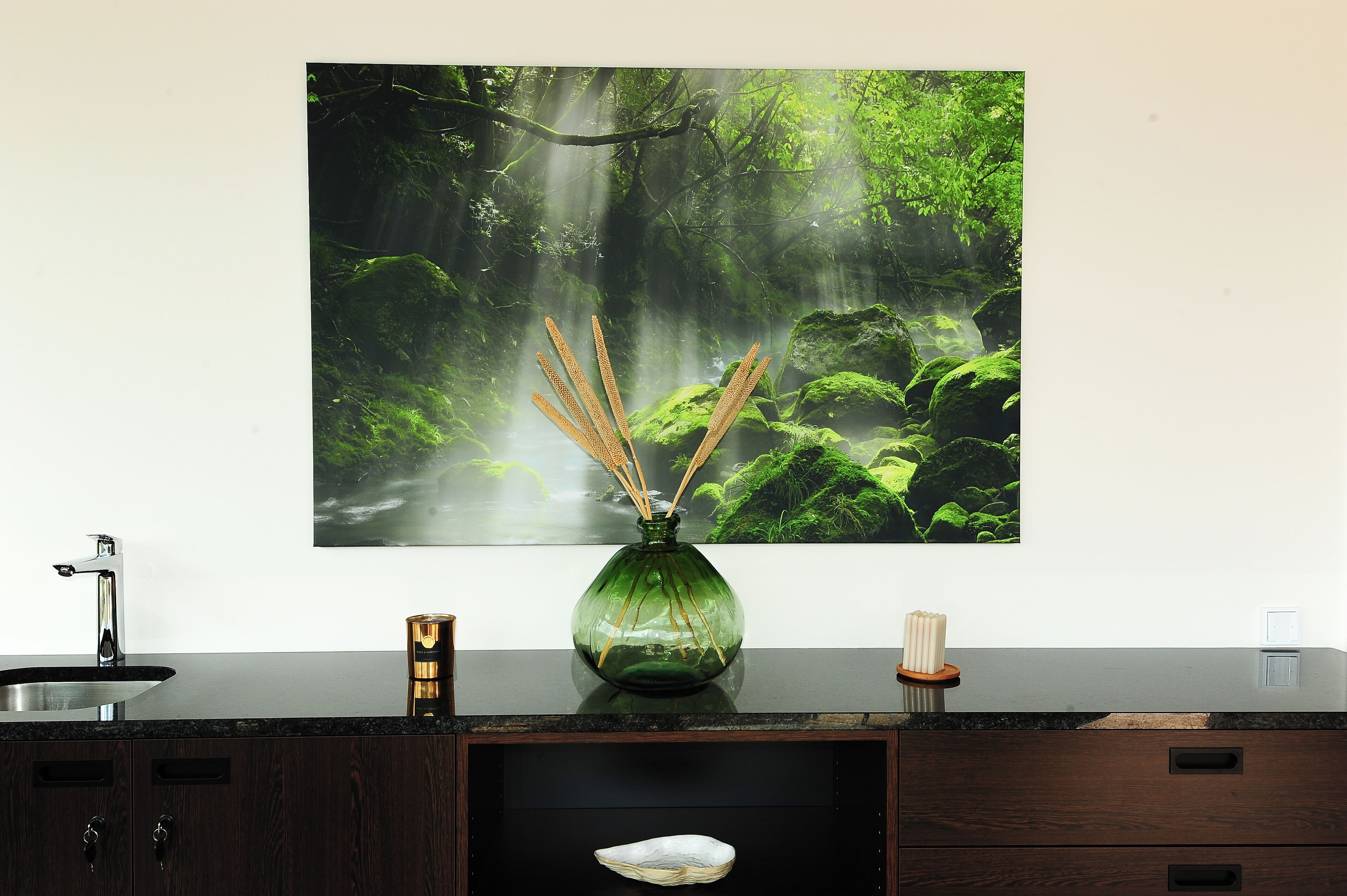 A bathroom with a sink, a green vase with reed sticks, and a large forest-themed poster on the wall.
