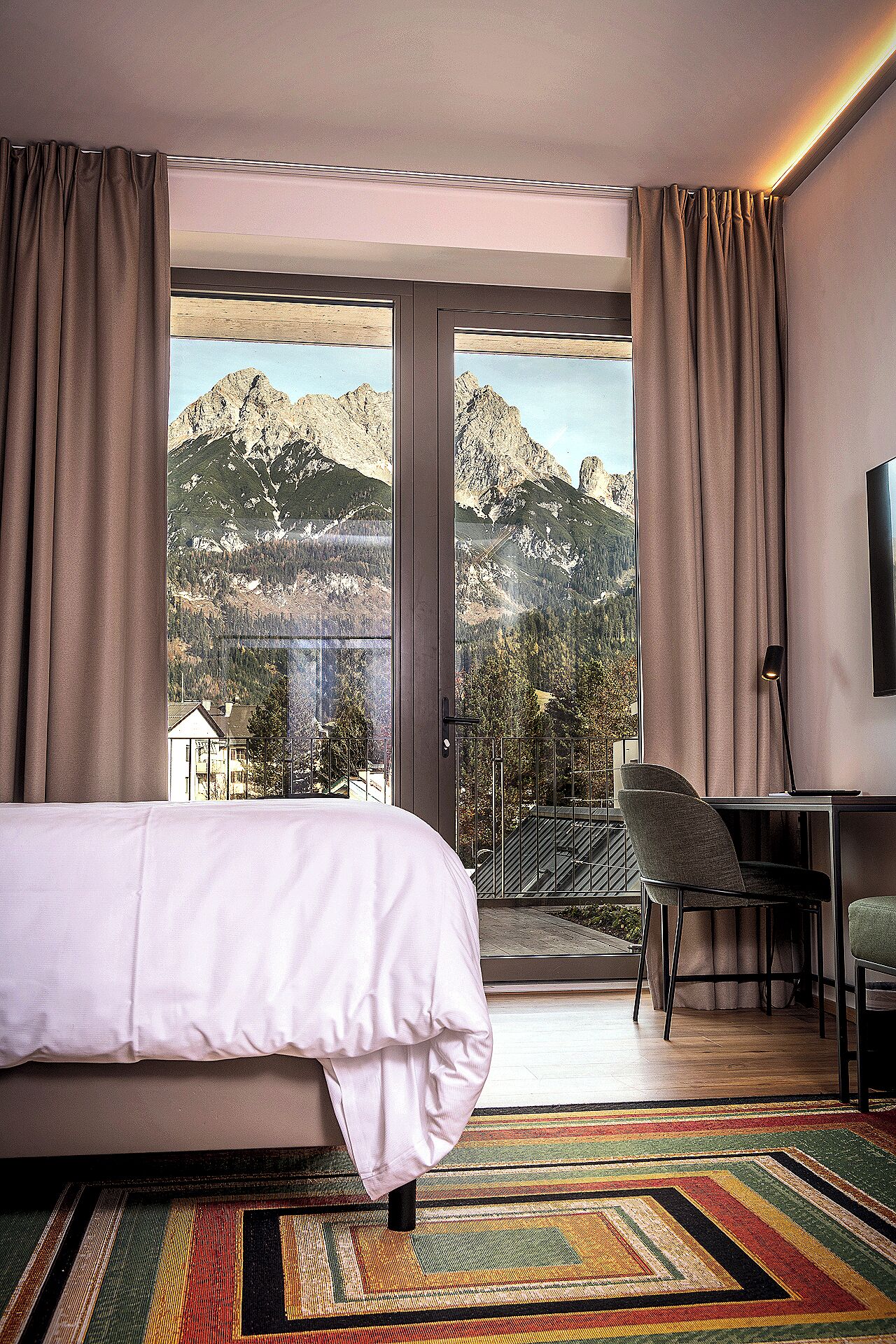A hotel room with a bed, a desk, a chair, and a large window offering a mountain view.
