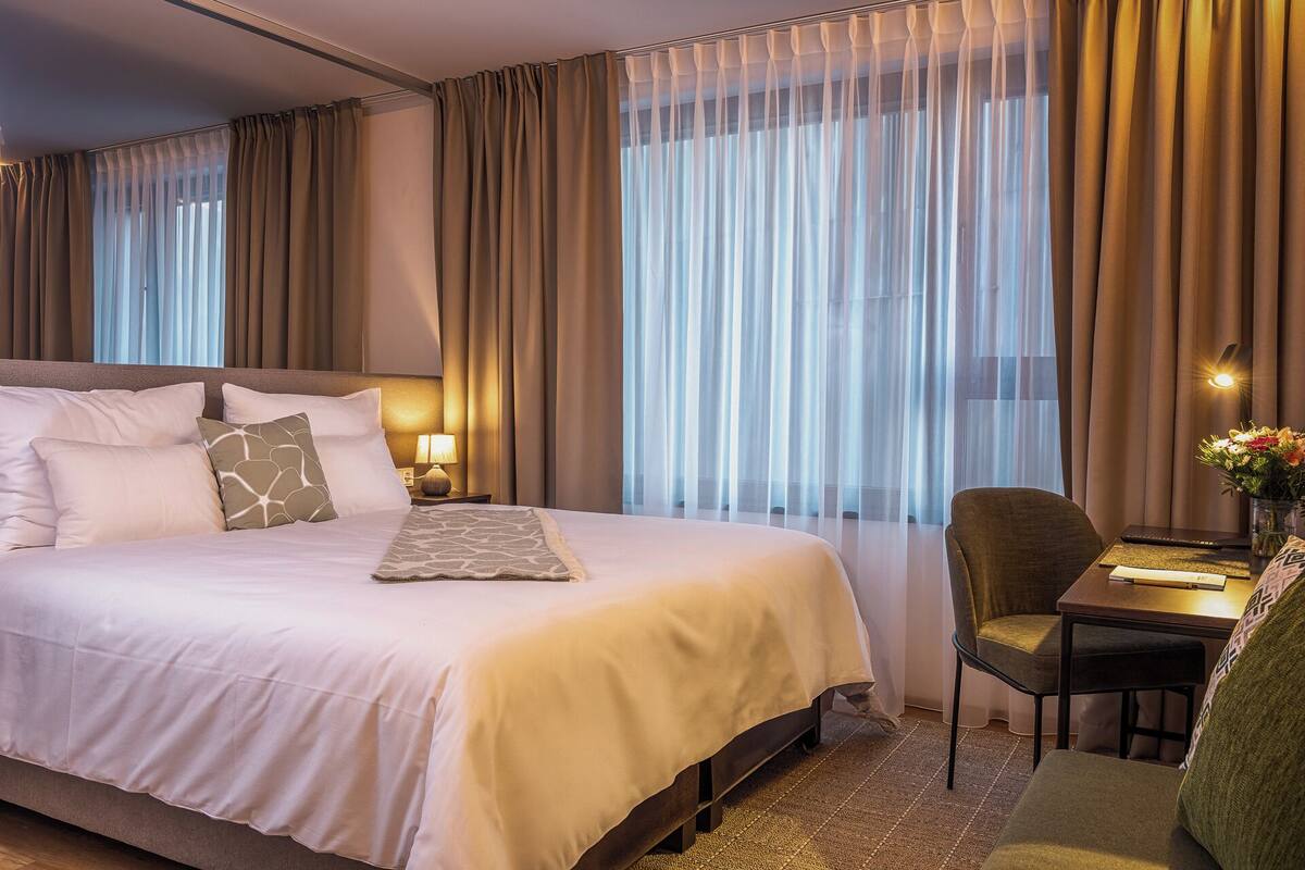 A hotel room with a large bed, a desk with a chair, and a window with curtains.