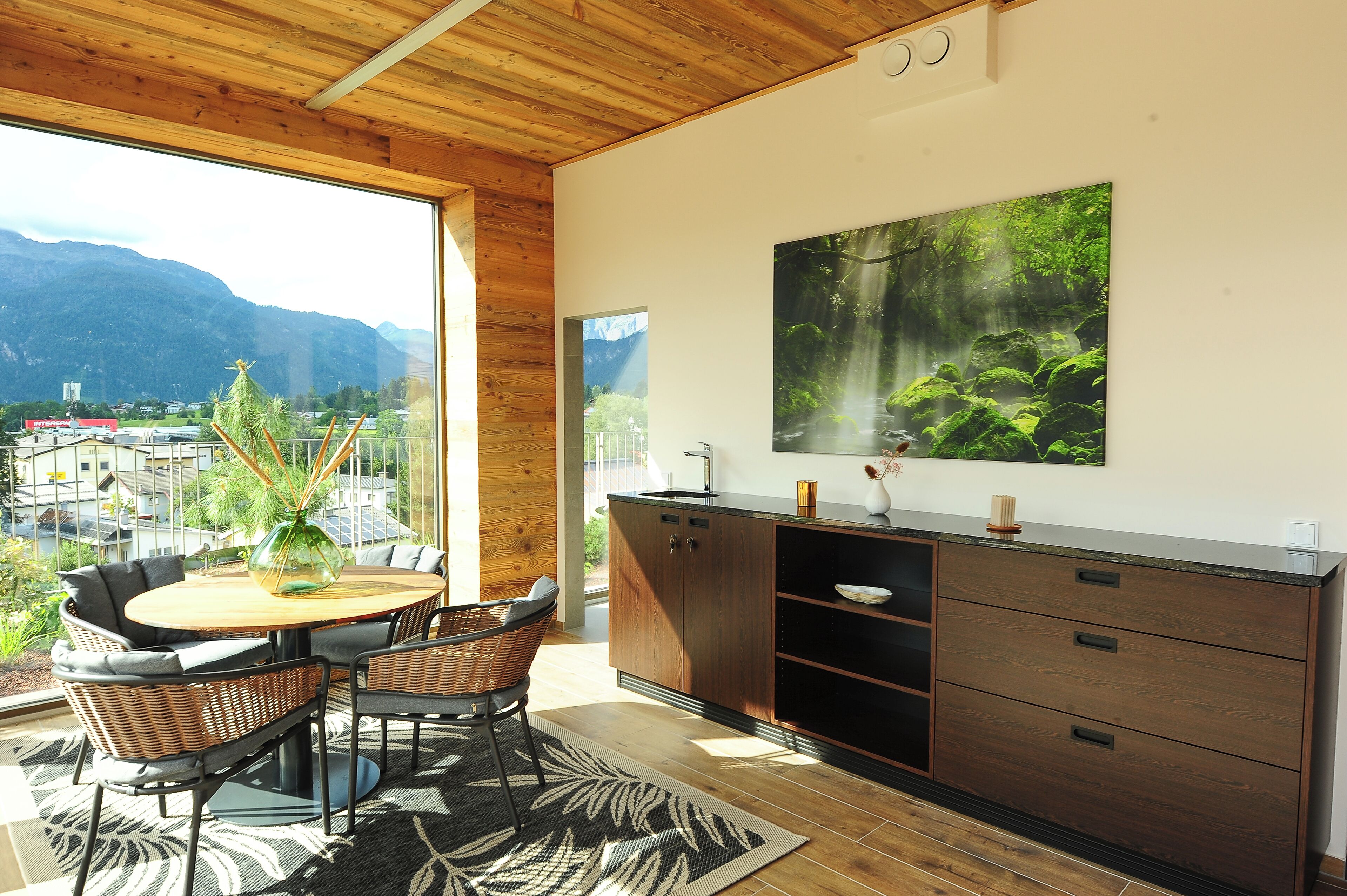 A modern kitchen with a large window, wooden ceiling, and a dining area with wicker chairs.