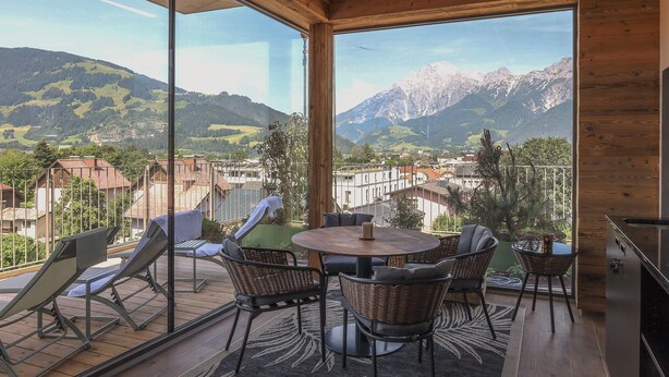 Un balcón con techo de madera, ventanas amplias, mobiliario de mimbre y vistas a montañas y un pueblo.