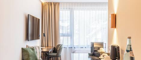 A modern hotel room with a wooden floor, a sofa with a green pillow, a dining table with chairs, a flat-screen TV mounted on the wall, a large window with sheer curtains, and a minibar with glasses and a bottle.