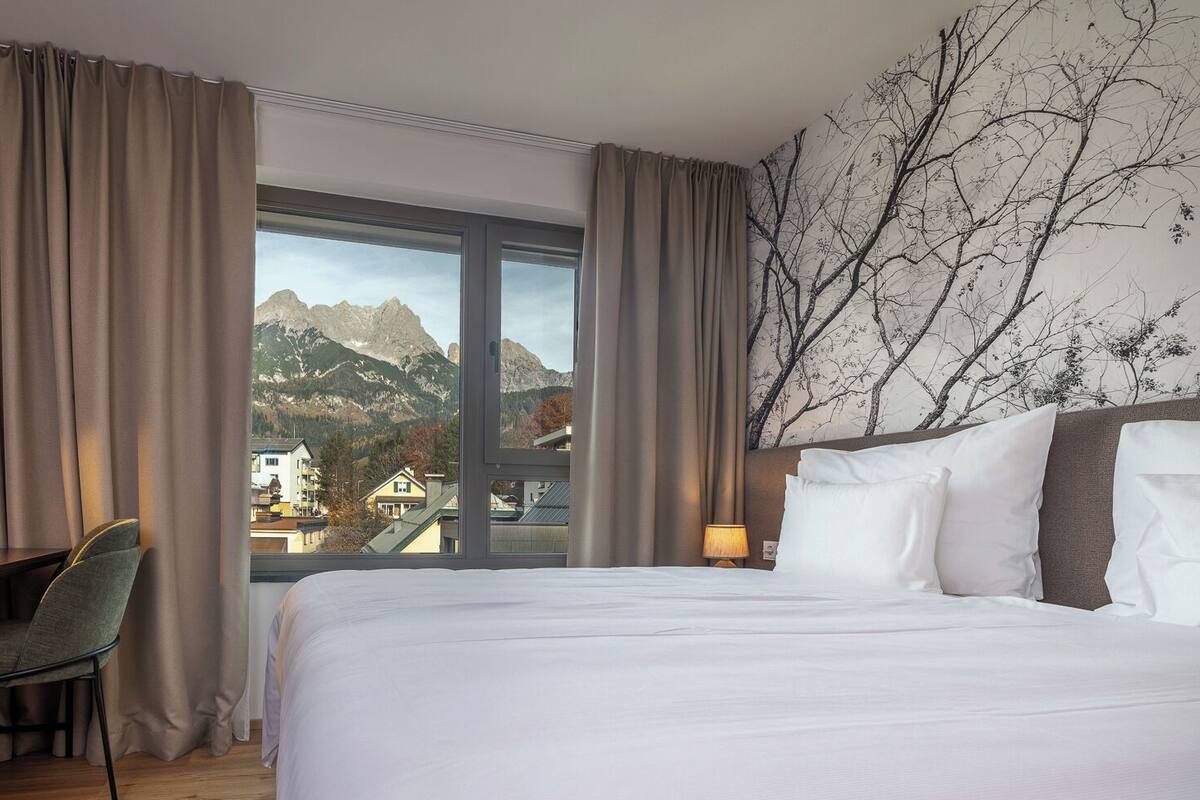 A hotel room with a bed, desk, chair, and a large window offering a mountain view.