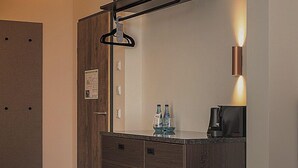 Una habitación de hotel moderna con un armario de madera, un perchero, una cafetera y una lámpara fijada a la pared.