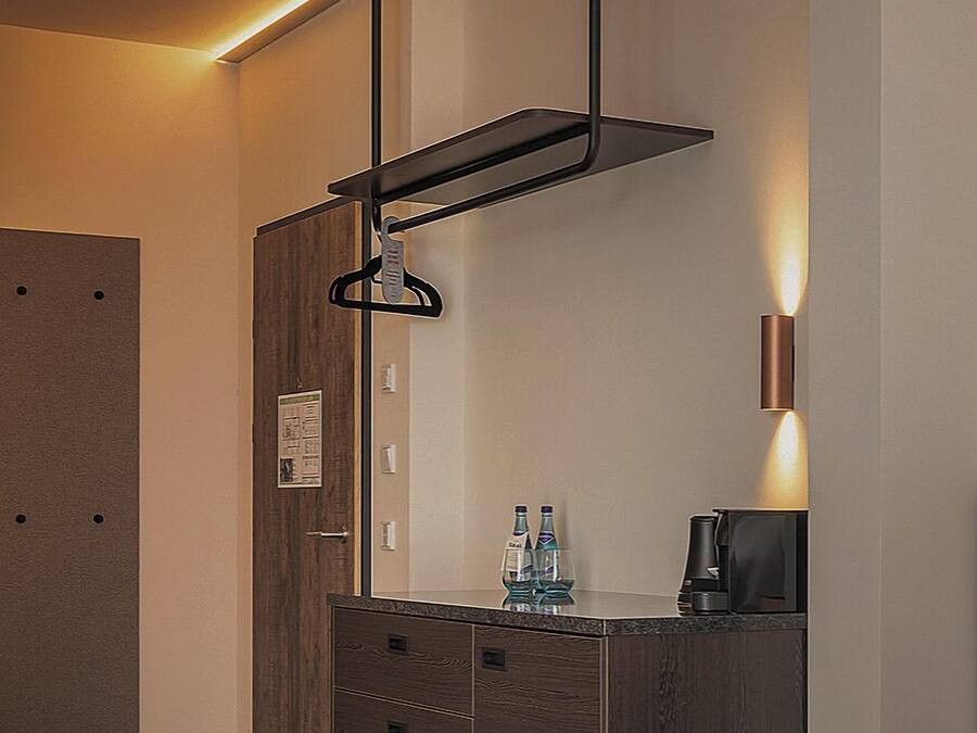 A modern hotel room with a wooden wardrobe, a hanging rack, a coffee machine, and a wall-mounted light.
