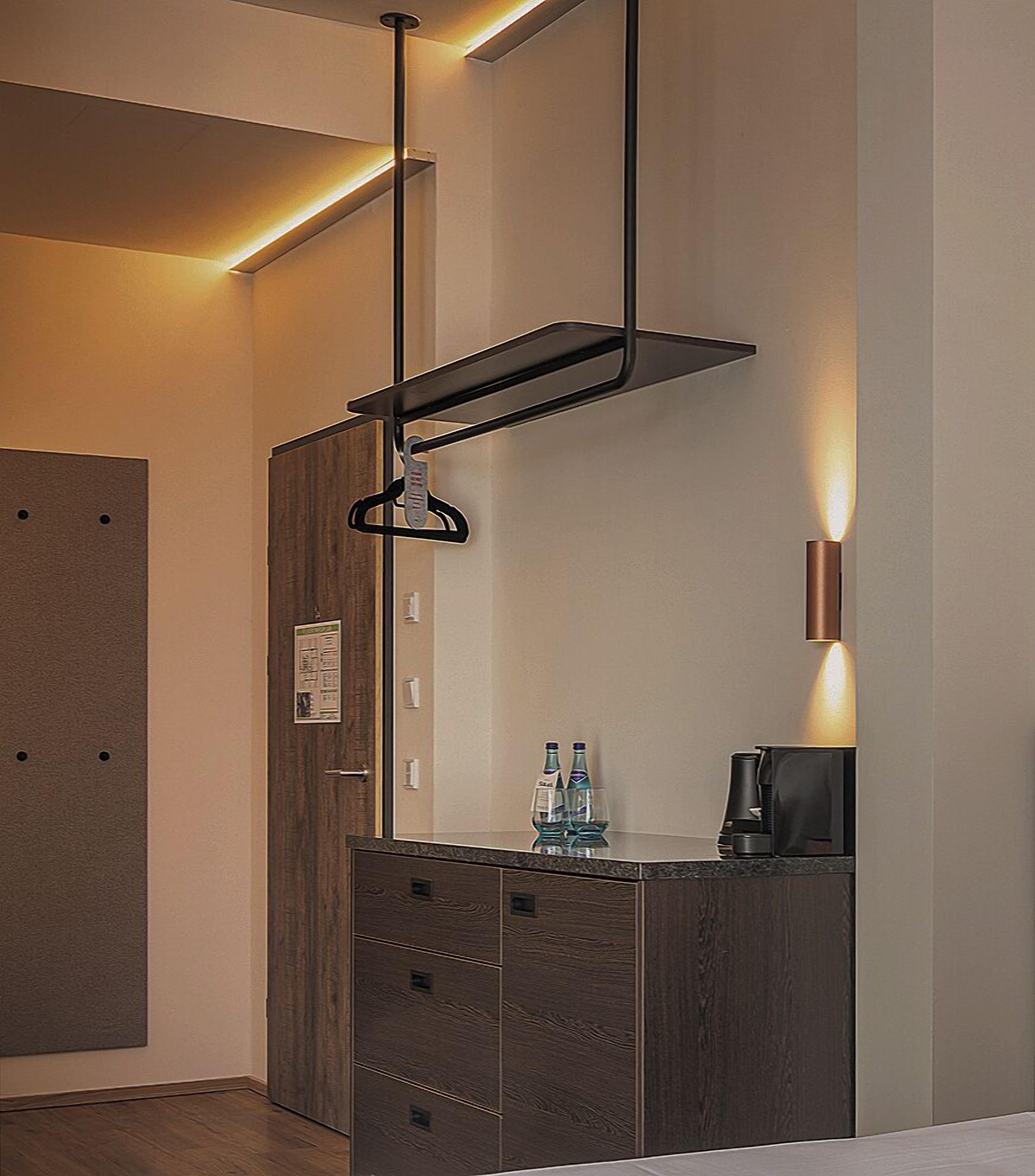 Una habitación de hotel moderna con un armario de madera, un perchero, una cafetera y una lámpara fijada a la pared.