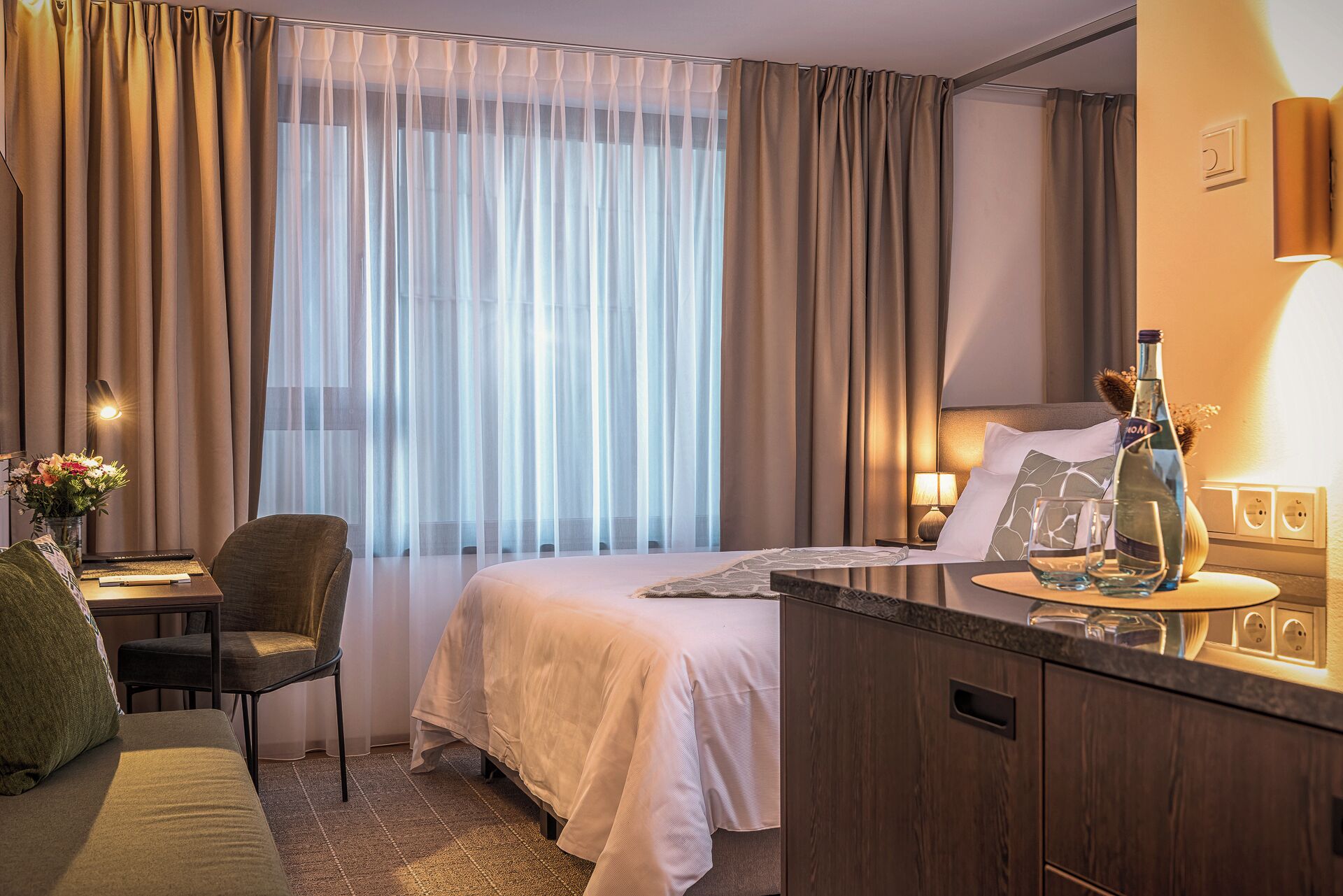 A hotel room with a bed, bedside table, lamp, and a desk with a chair.