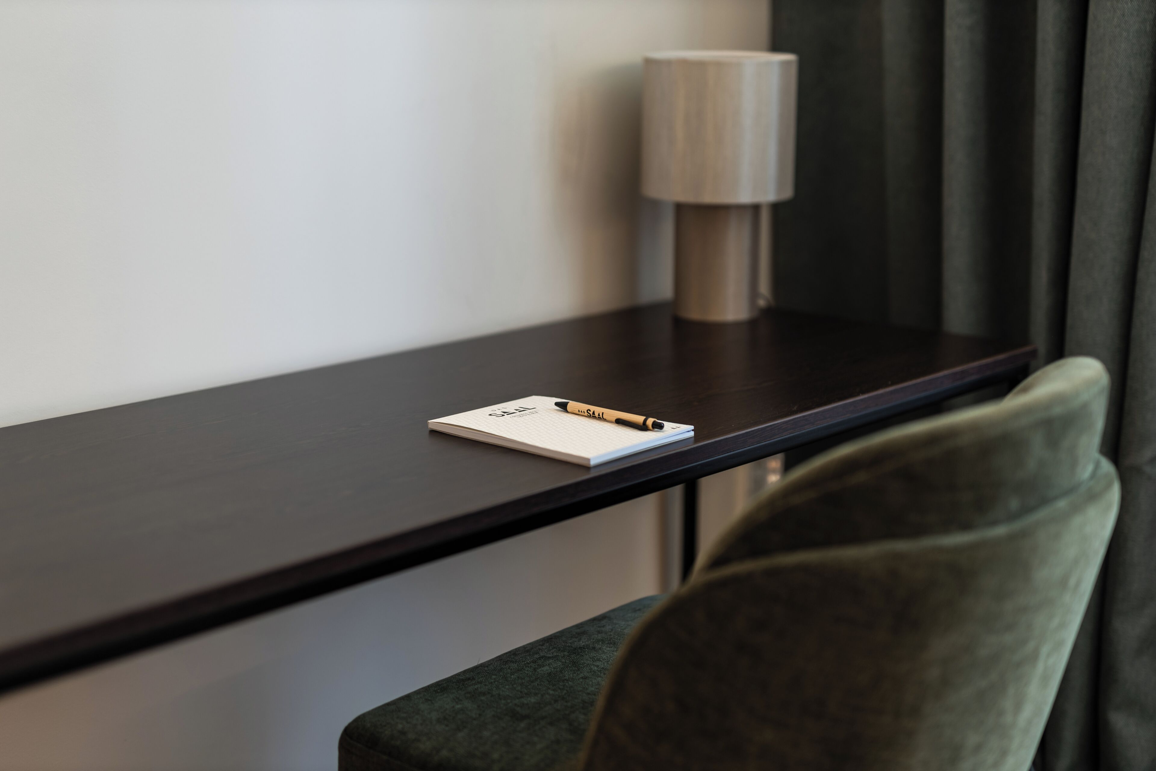 A neatly arranged hotel room with a dark wooden desk, a lamp, a notepad, and a pen.