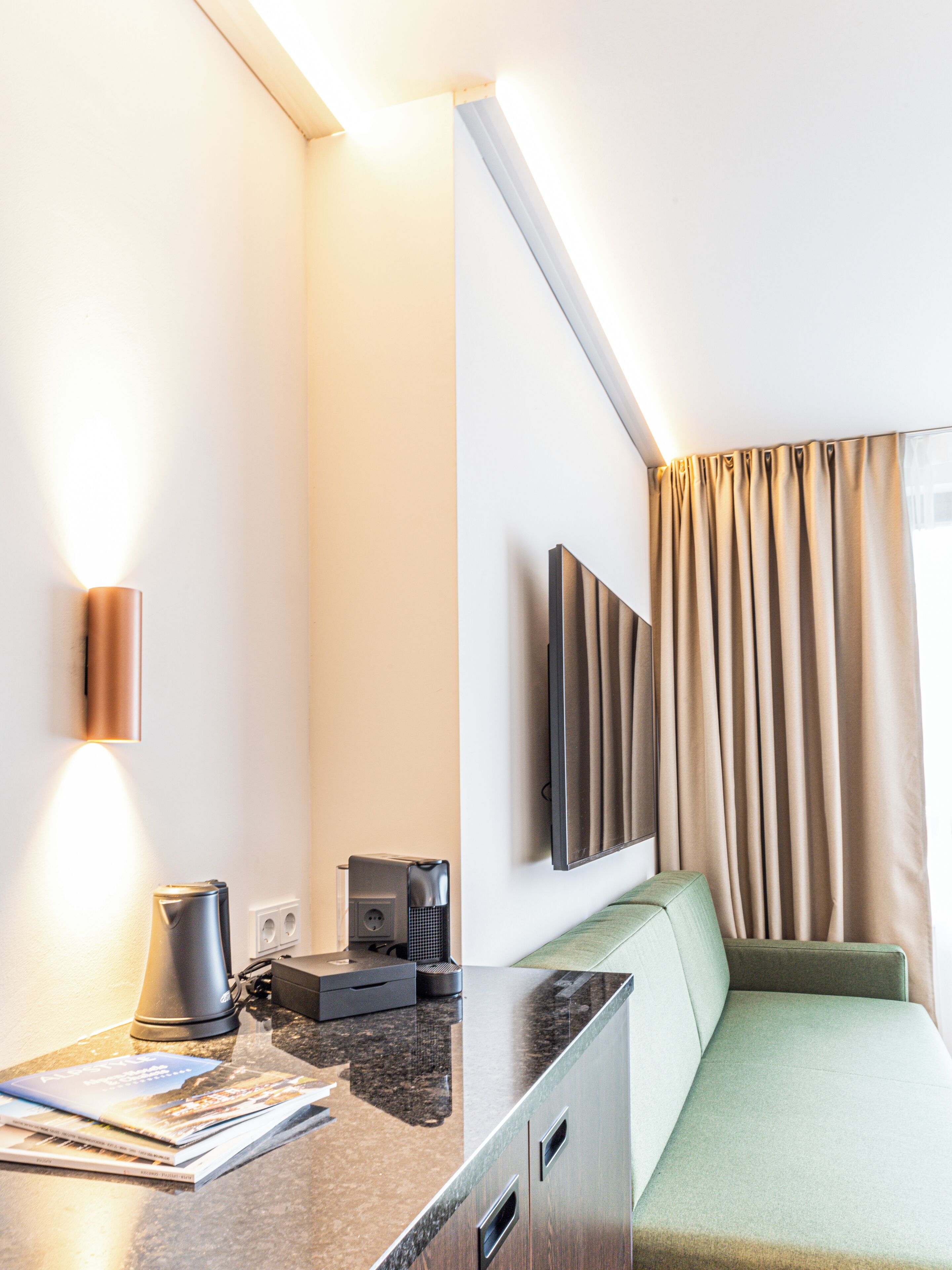 A modern hotel room with a flat-screen TV mounted on the wall, a sofa, a coffee machine, and a countertop with a magazine.
