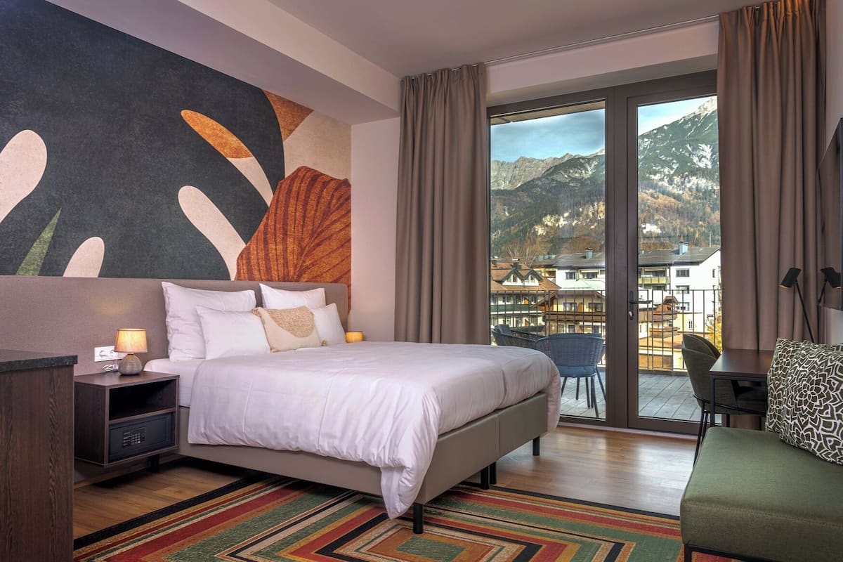 A hotel room with a large bed, a desk, a chair, and a balcony with a mountain view.