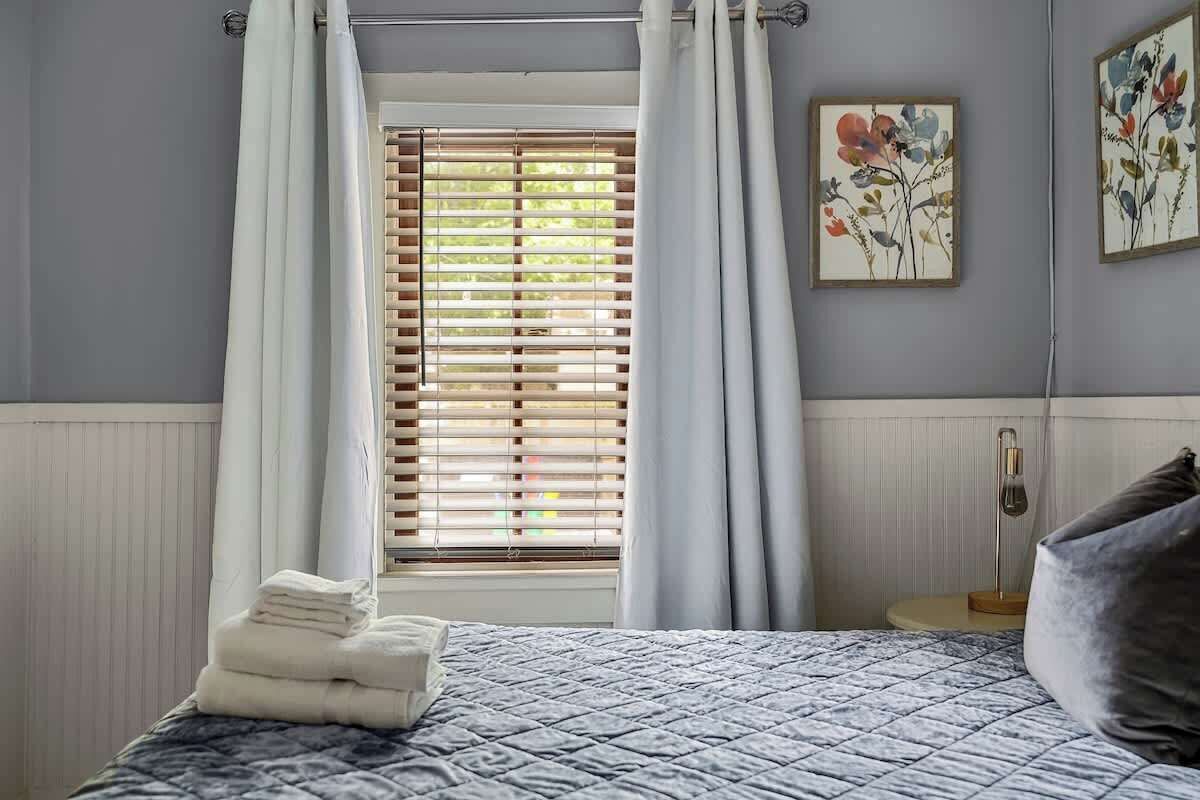 Un dormitorio con cama, ventana con persianas, mesita de noche con lámpara y dos cuadros con motivos florales en la pared.