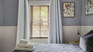 A bedroom with a bed, a window with blinds, a bedside table with a lamp, and two floral paintings on the wall.