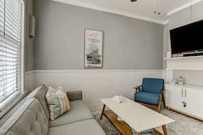 A living room with a grey sofa, a blue armchair, a white coffee table, and a flat-screen TV on a white cabinet. - Hot Tub - Historic District - Next to Forsyth Park (Savannah)