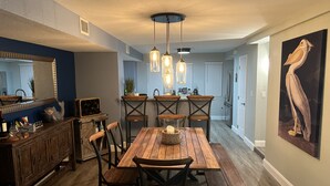 A dining area with a wooden table and chairs, a kitchen in the background, and a large painting on the wall.