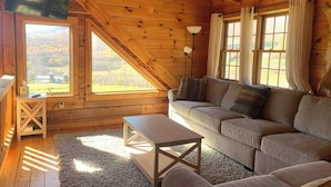 Ein gemütliches Blockhaus-Interieur mit Holzbalkendecke, einer Couch, einem Tisch und großen Fenstern mit Blick ins Freie.