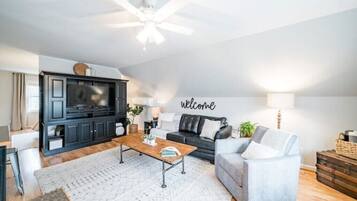 Un salon moderne avec un meuble de rangement noir pour l’équipement audiovisuel, un canapé gris, une table basse en bois et un panneau mural portant l’inscription « Bienvenue ».