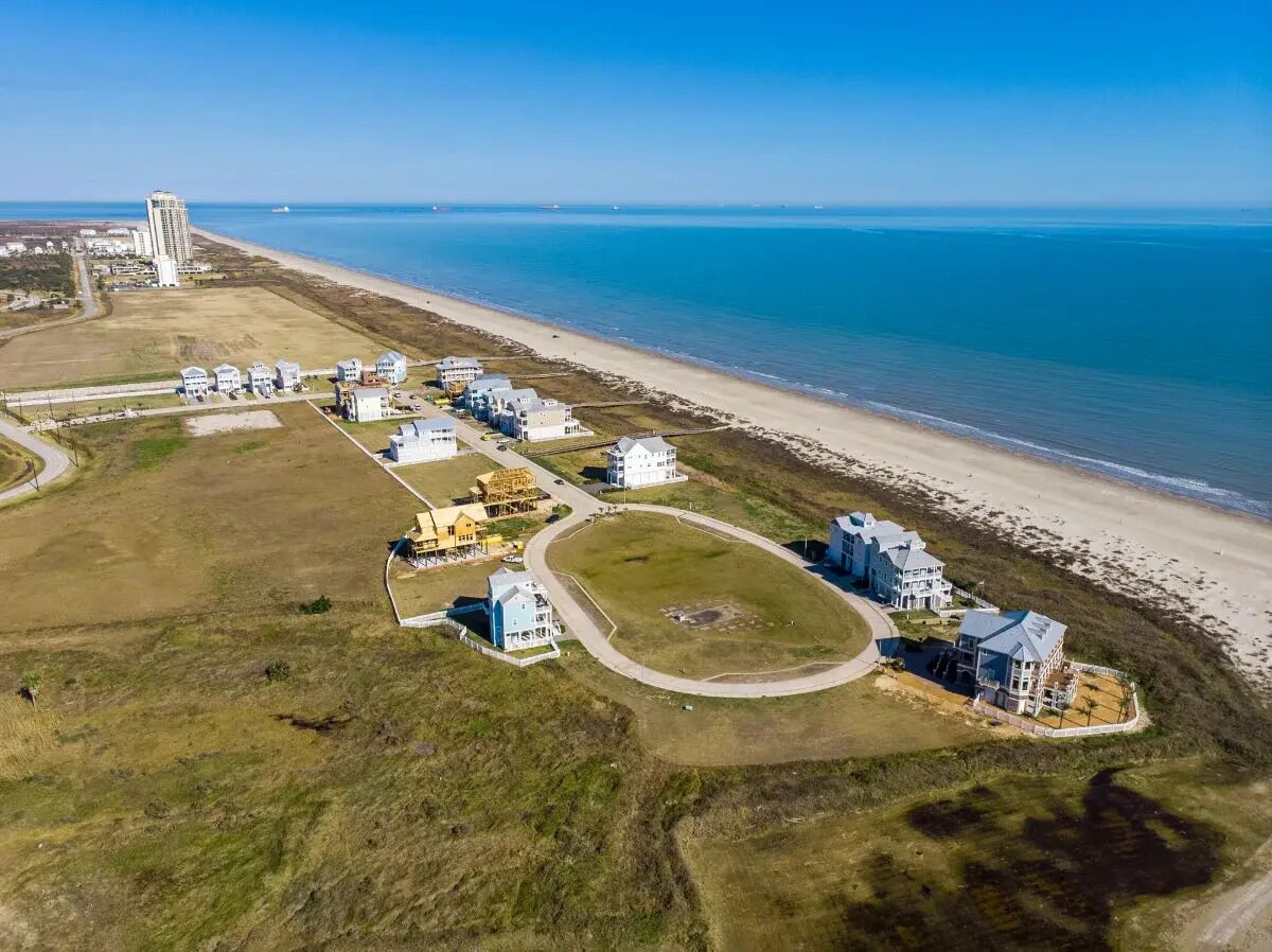 Vista aérea de una zona residencial costera con casas cerca de la playa, un edificio grande a lo lejos y un cielo azul despejado.