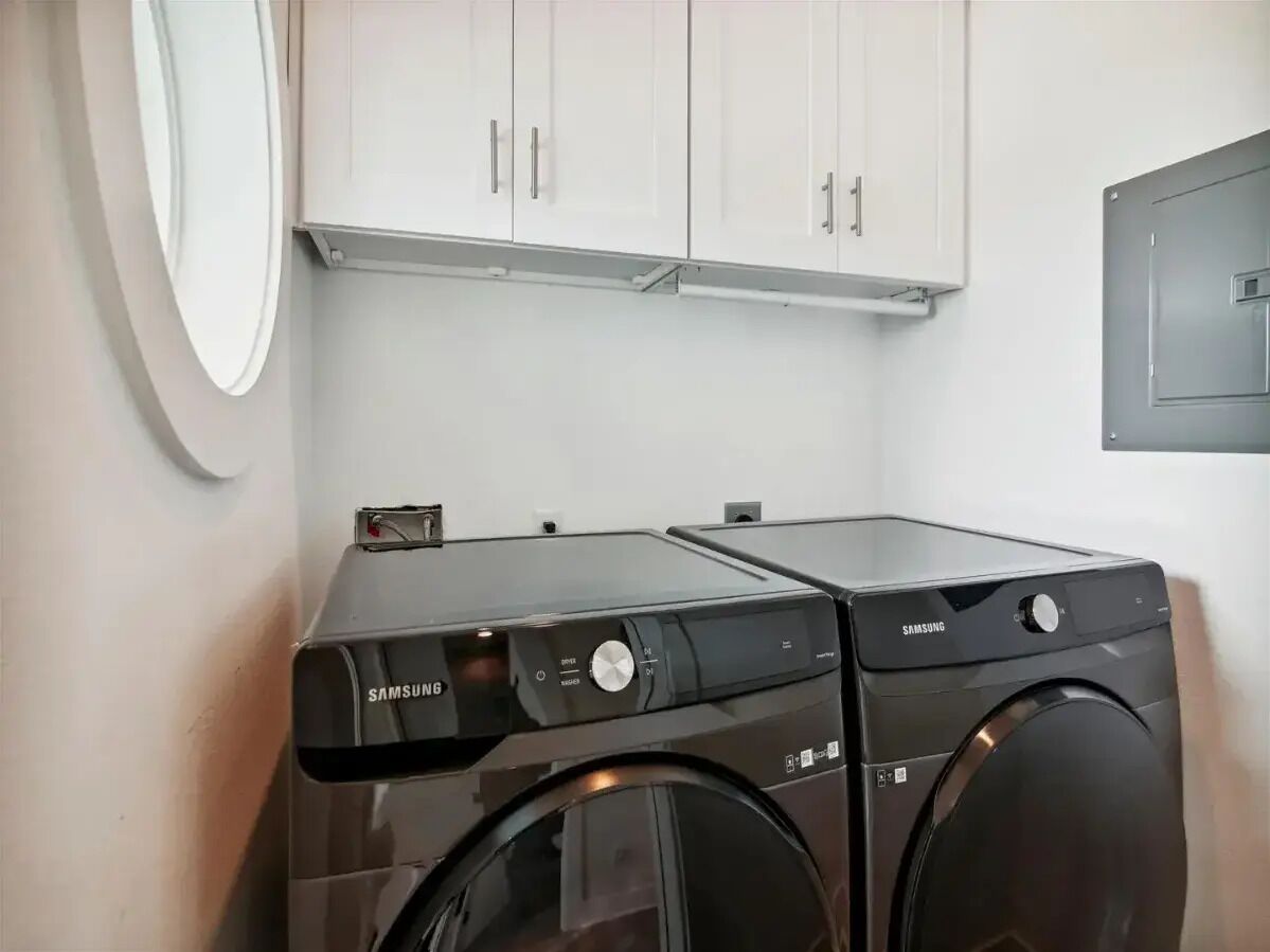 A laundry area with two Samsung washing machines, a white cabinet above, and a wall-mounted electrical box.