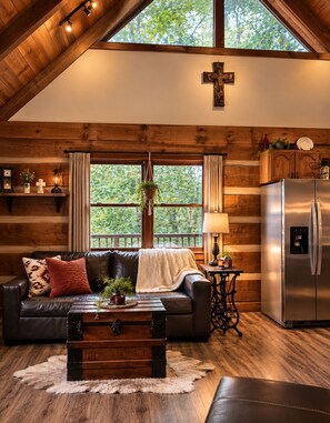 A cozy cabin interior with a wooden ceiling, a cross on the wall, a sofa with cushions, a coffee table, a refrigerator, and a window with curtains.