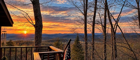 A wooden deck with an American flag, overlooking a forested landscape with a sunset.