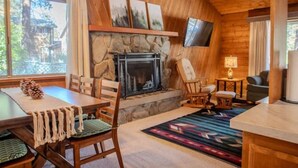 A cozy cabin interior with a stone fireplace, wooden walls, a dining area, and a television mounted on the wall. - Pinetree Place (Wrightwood)