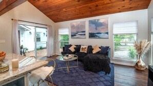 Ein gemütliches Wohnzimmer mit einem dunkelblauen Teppich, einer schwarzen Couch und einem Glastisch. An der Wand hängen drei gerahmte Bilder und es gibt ein Fenster mit Jalousien.