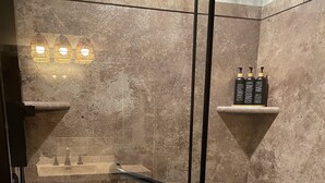 Un área de ducha moderna con puerta de vidrio, una silla de madera y un estante con artículos de aseo.