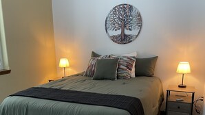 Un dormitorio ordenado con una cama, dos mesitas de noche, una lámpara de pared y un cuadro con un árbol decorativo.