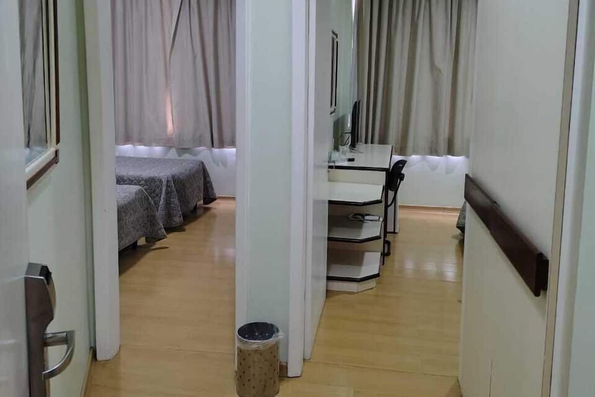 Una habitación de hotel con piso de madera, una cama con cubrecama gris, un escritorio con silla y un basurero pequeño.