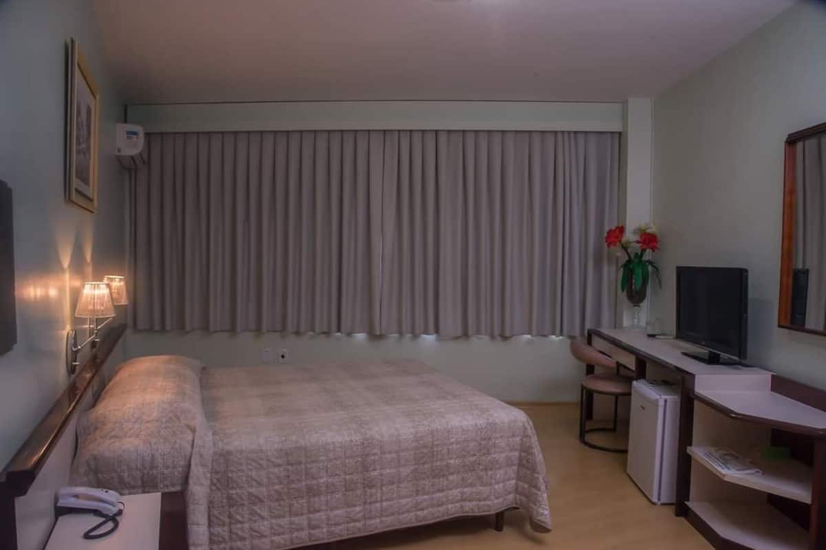 Habitación de hotel con cama, mesita de noche con lámpara, escritorio con computadora, televisor y ventana con cortinas.