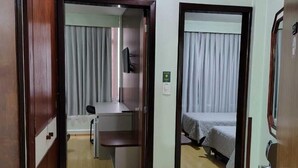 Ein Hotelzimmer mit Holzboden, einem Schreibtisch mit Stuhl, einem Bett mit grauer Bettdecke und einem kleinen Mülleimer.