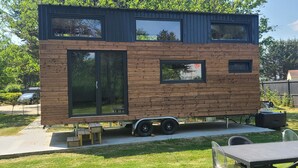 A modern, two-story mobile home with large windows and a flat roof, situated in a grassy backyard.